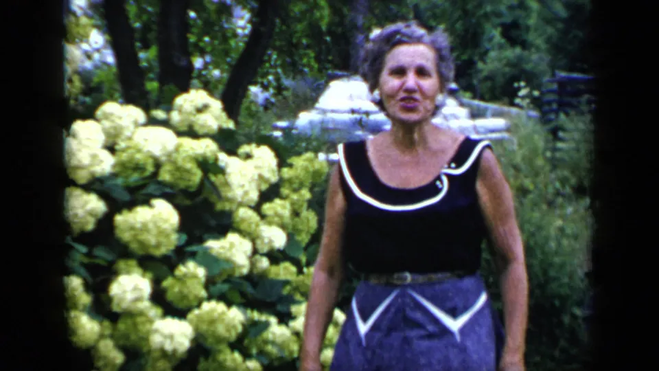 The woman remains near blooming yellow shrubs in the garden, with trees and soft 8mm color grain behind her.