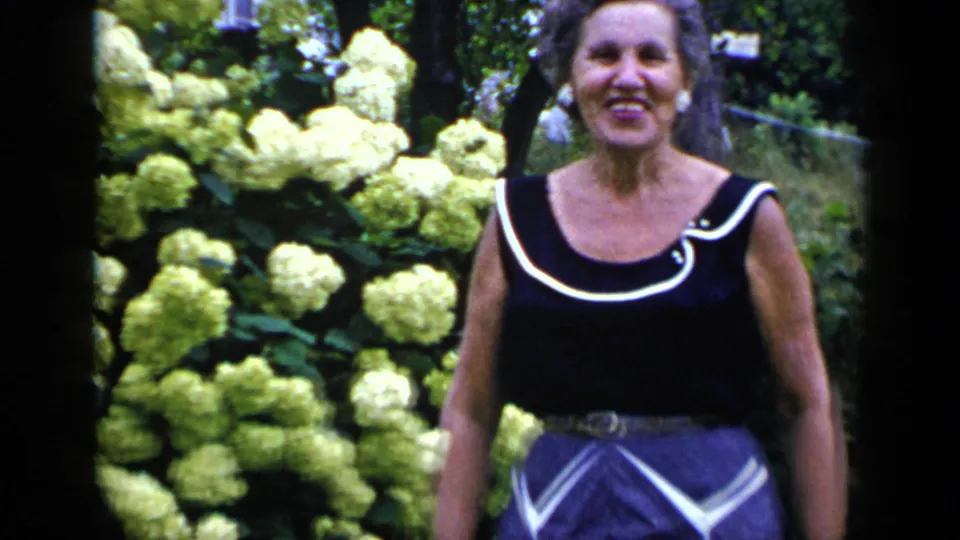 Smiling adult woman in a black dress with white trim stands near pale hydrangeas in a 1948 New York garden scene.