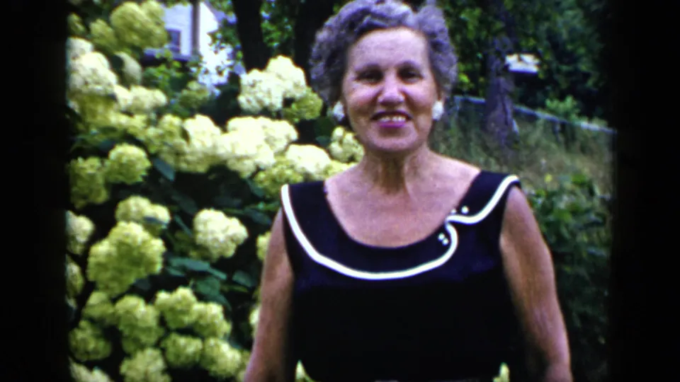 Adult woman with short curled hair stands closer to the camera, framed by pale hydrangeas and garden greenery in New York.