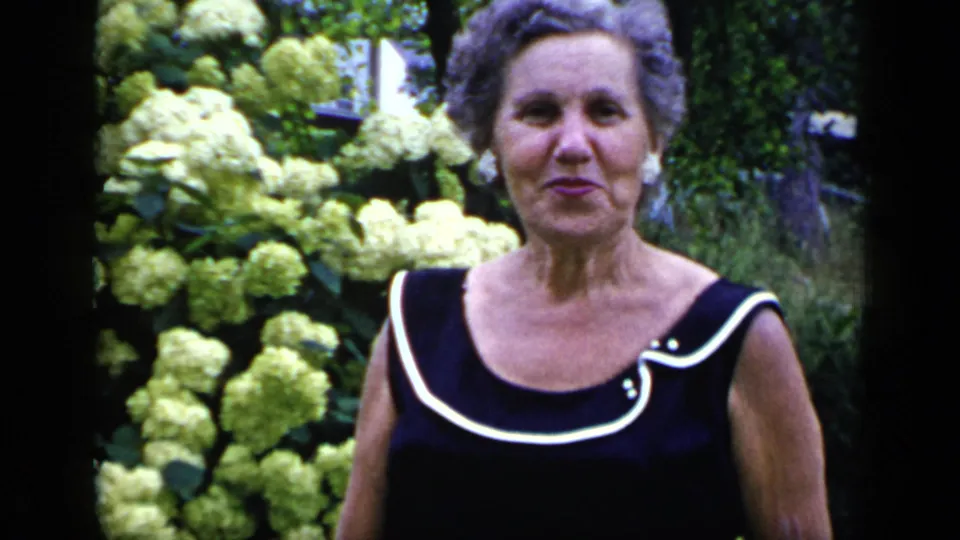 Adult woman in a black sleeveless dress with white trim looks toward the camera, with pale hydrangeas and foliage behind her.
