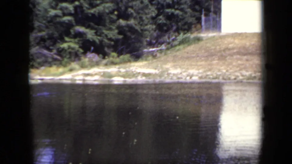 1948 New York 8mm frame showing a pond, grassy bank, dense trees, and a white rectangular structure reflected in the water.