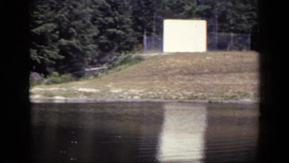 1948 New York 8mm view of a pond shore with a white rectangular structure, tree line, grassy slope, and soft water reflection.