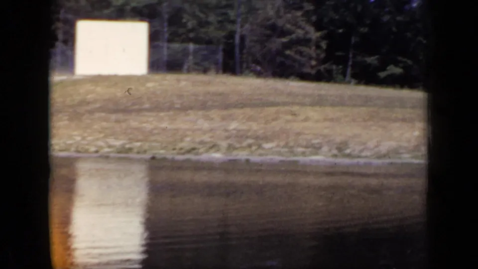 1948 New York 8mm frame with the pond lower in view, a grassy shore, trees, and a white rectangular structure near the left side.