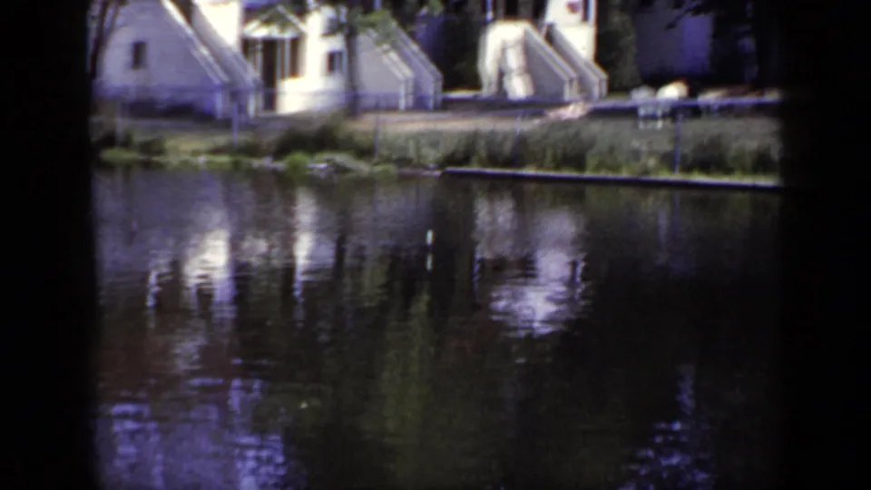 1948 New York 8mm frame showing pond reflections, small residential houses, exterior steps, trees, and shoreline fencing.