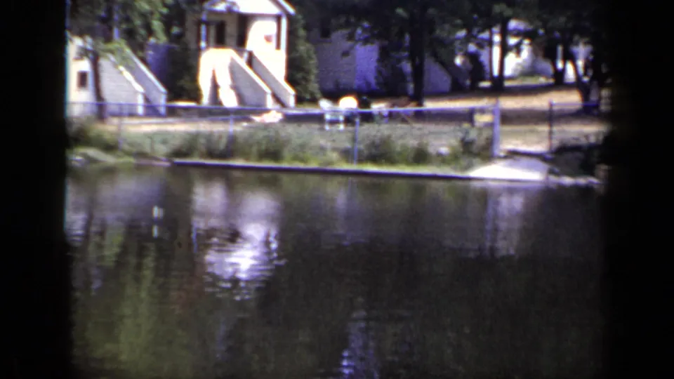 1948 New York 8mm view across a pond toward small houses, chain-link fencing, trees, and reflected light on the water.