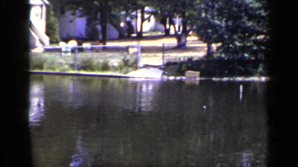 1948 New York 8mm frame showing a pond edge with trees, chain-link fence, walkway, shoreline plants, and a small dock.