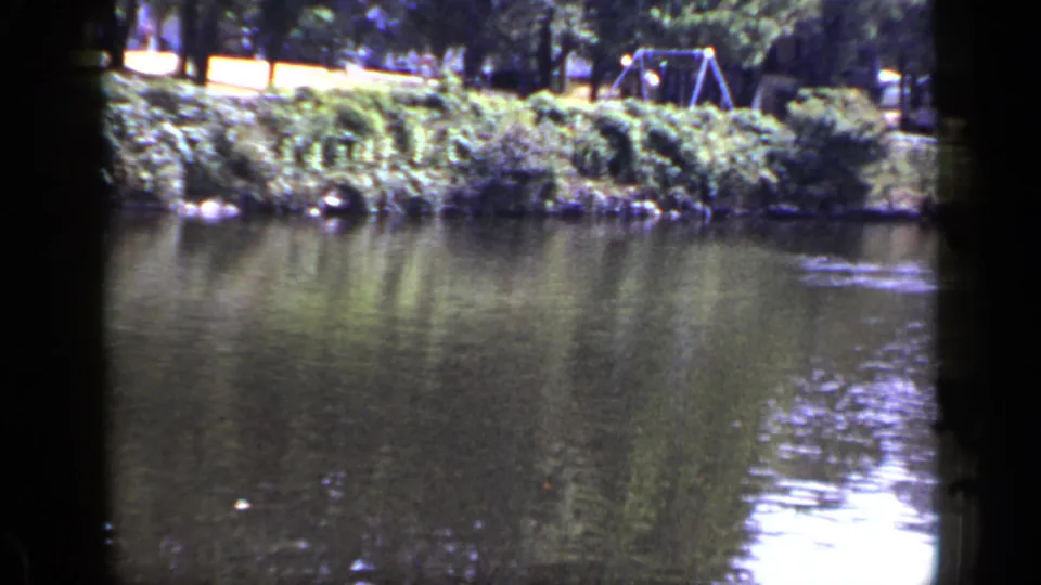 1948 New York 8mm frame showing pond water, shrubs, trees, and a small playground structure in the distance.