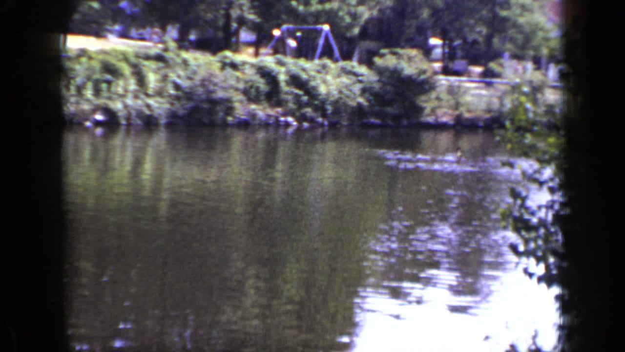 1948 New York 8mm frame looking across a calm lake toward trees, grass, and a swing set on the far bank.