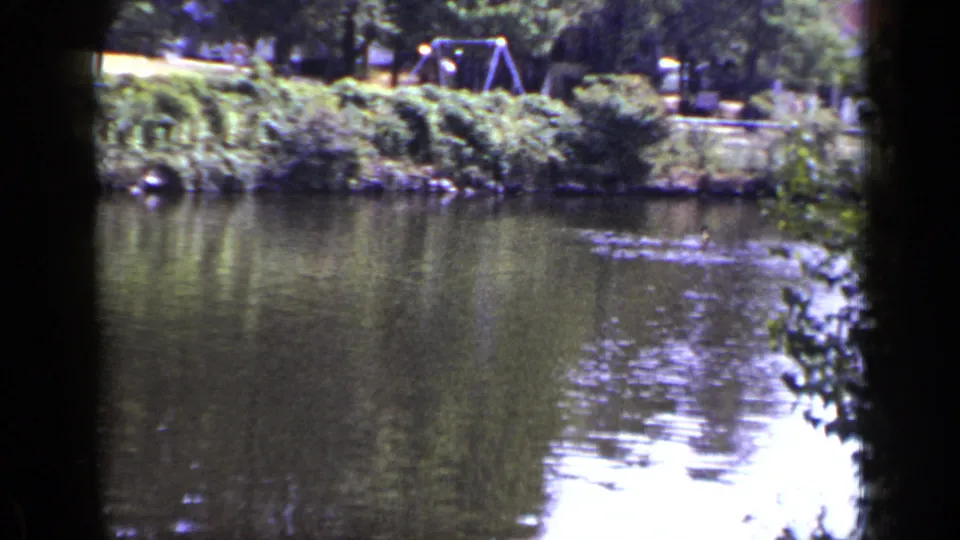 1948 New York 8mm frame looking across a calm lake toward trees, grass, and a swing set on the far bank.