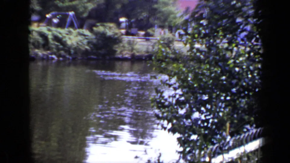 1948 New York home-movie frame with lake water, far-bank greenery, and dark gate-edge framing as the camera pans.