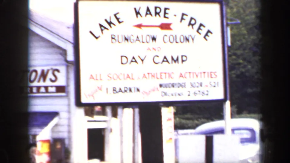 1948 New York frame showing the Lake Kare-Free Bungalow Colony and Day Camp sign with building edge, fence, and parked vehicle nearby.