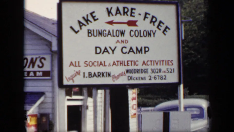 1948 New York 8mm frame showing a large Lake Kare-Free Bungalow Colony and Day Camp sign with contact details near a roadside building.