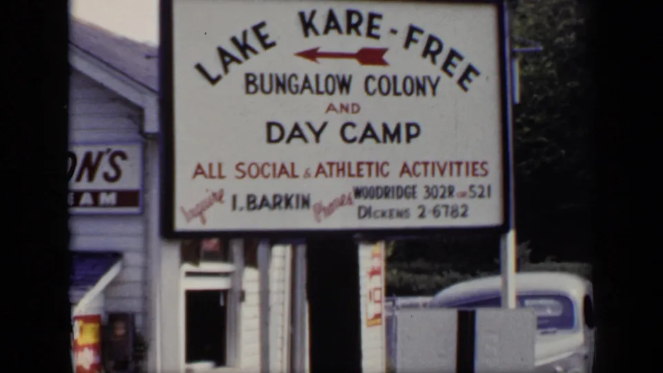 1948 New York frame of the Lake Kare-Free Bungalow Colony and Day Camp sign, with phone details and a partial building behind it.