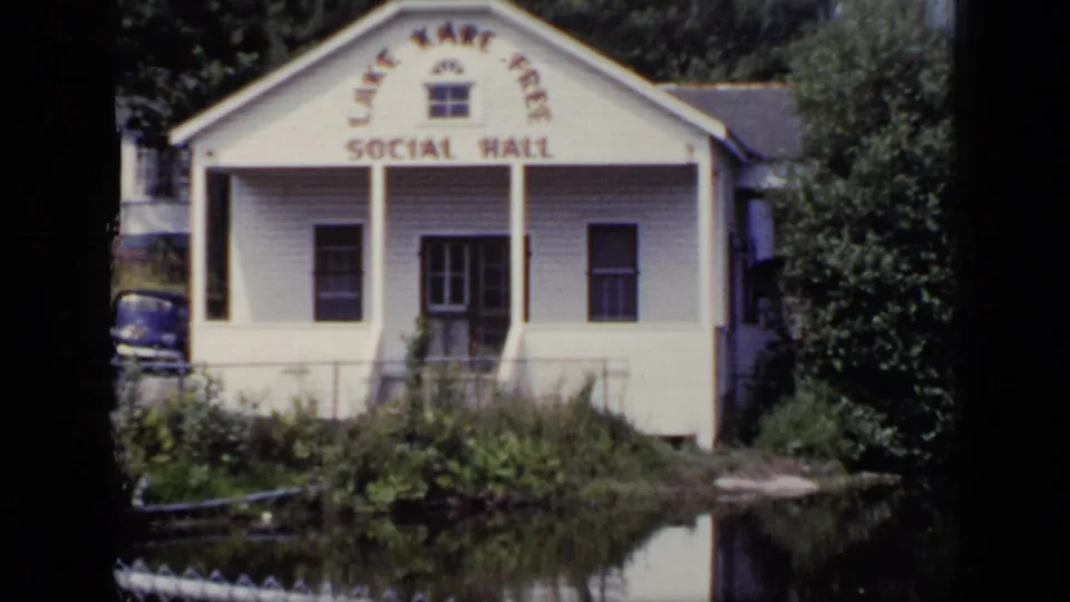 1948 New York frame showing the Lake Kare Free Social Hall, a white clapboard building by water with trees and brush around it.