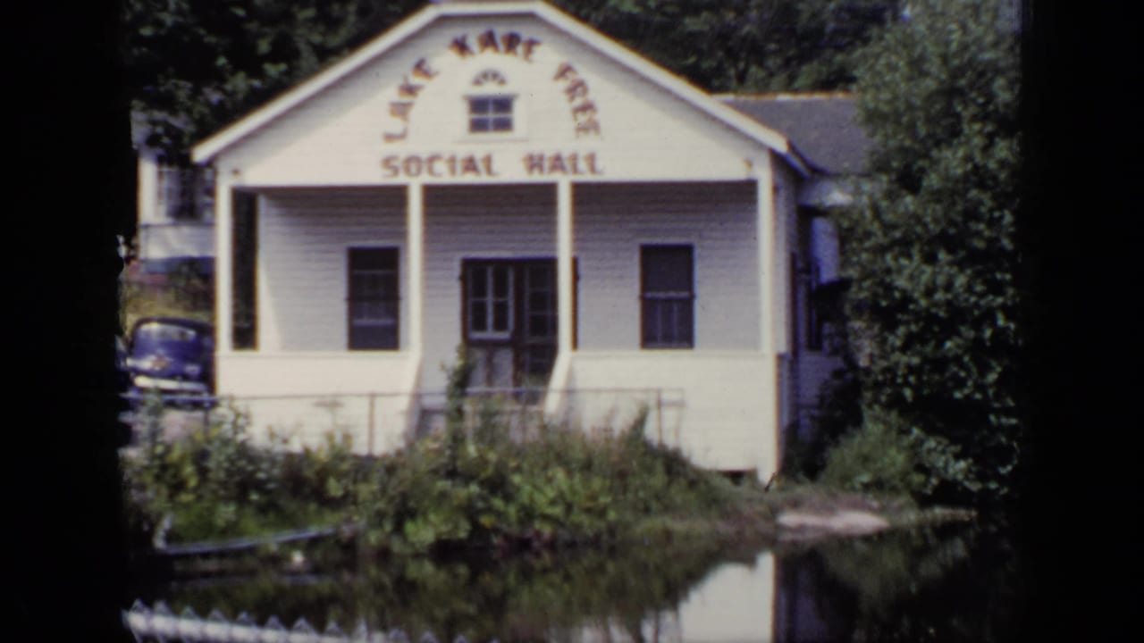 White social hall across a pond with trees and parked cars nearby.