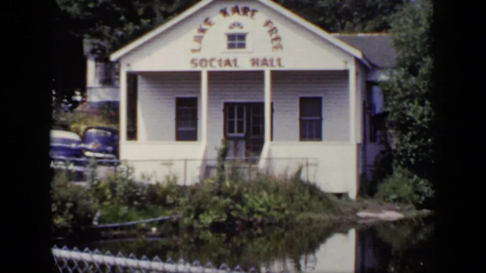 White Lake Kari Free Social Hall beside pond water, with mid-century cars and trees visible in the New York scene.