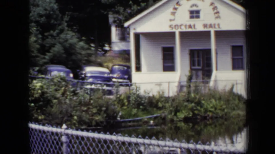 Closer 1948 home-movie view of the Lake Kari Free Social Hall, parked cars, chain-link fence, and pond edge.