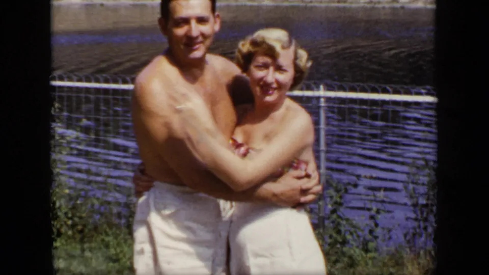 Adult man and woman in 1940s-style bathing suits embrace by pond water in the New York home-movie footage.