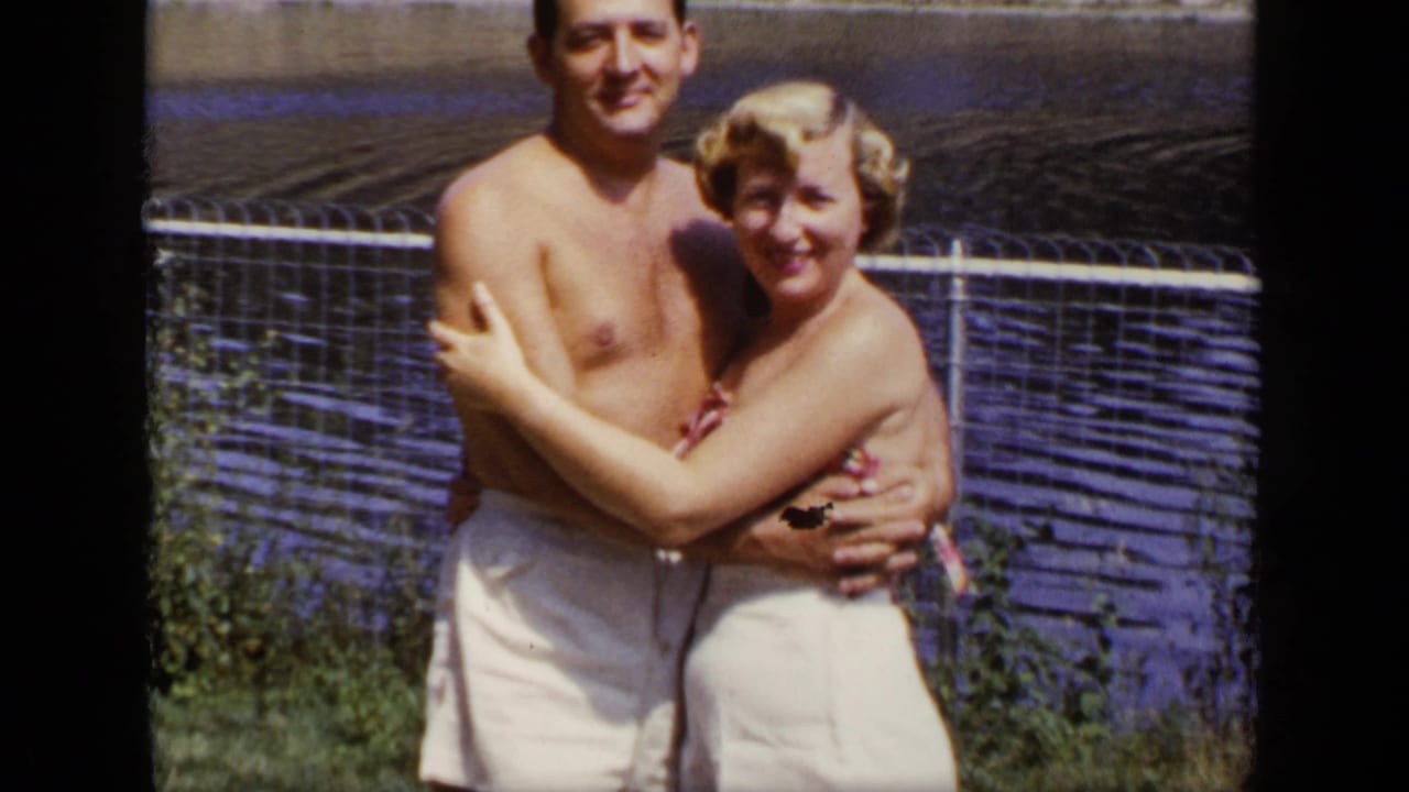 In 1948 New York home-movie footage, a young couple in swimwear stands arm in arm by a chain-link fence near lake water.
