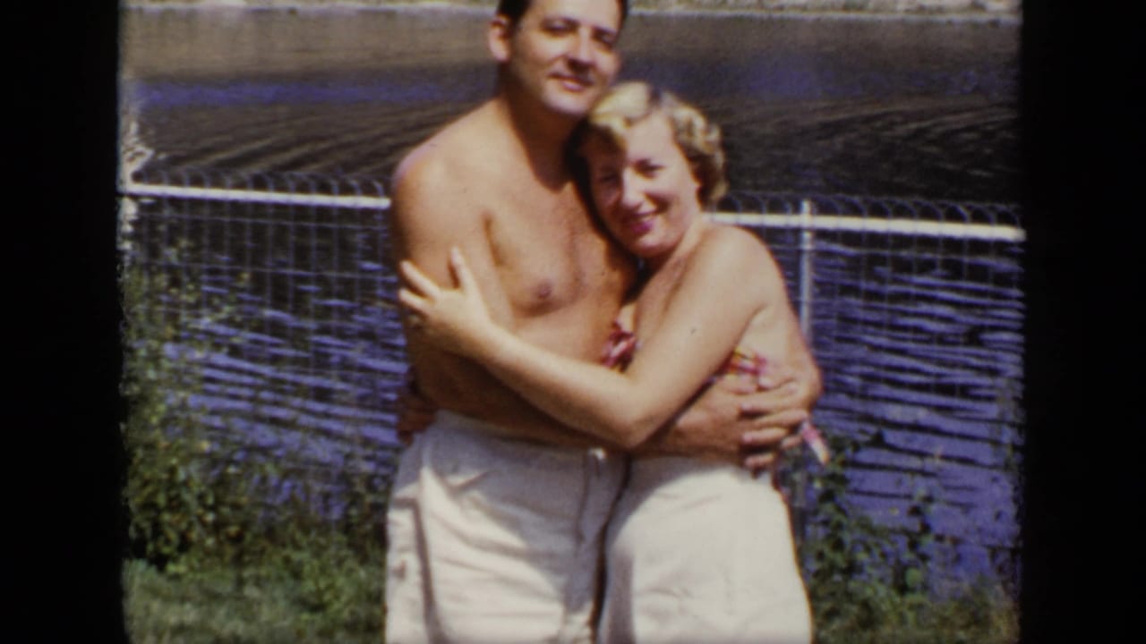 Couple poses closely by a chain-link fence with lake water behind them.