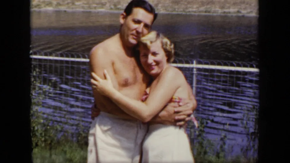 A young woman in a light swimsuit leans into a shirtless man as they pose by a fence and lake in the New York home movie.