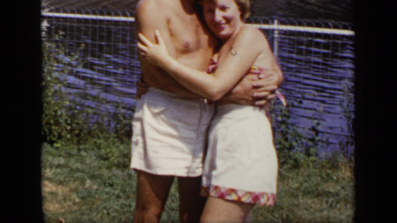 Adult couple embraces on grass beside water and fencing.