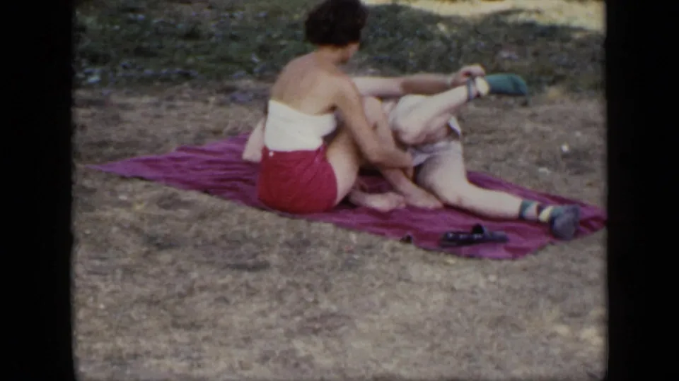 An adult couple relaxes on a red blanket in a grassy outdoor area, with the woman seated upright and the man reclining.