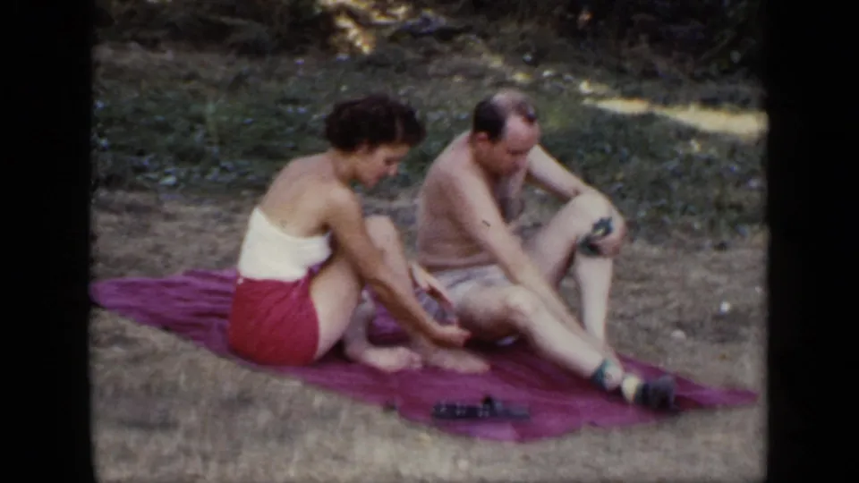 The woman sits beside the shirtless man on a purple blanket as he leans forward in a sun-dappled New York park setting.