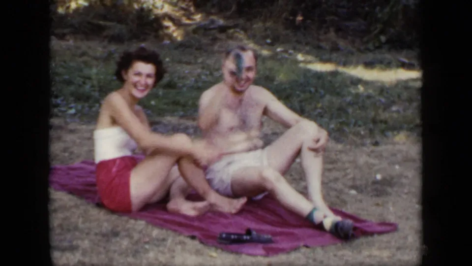 The seated couple smiles at the camera while the shirtless man raises a small object during a 1948 New York park home movie.