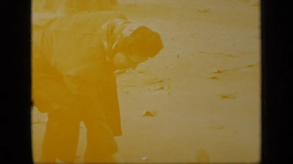 1948 New York home-movie frame with an older woman in a coat and patterned scarf bending over on pavement with autumn leaves nearby.