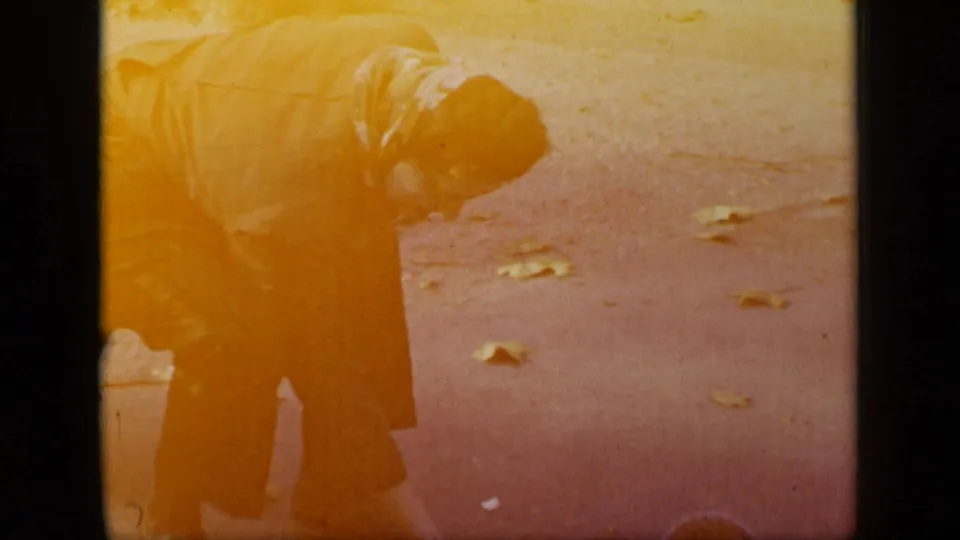 Warm yellow 8mm frame from the New York clip showing the woman bending close to the pavement as autumn leaves remain visible around her.