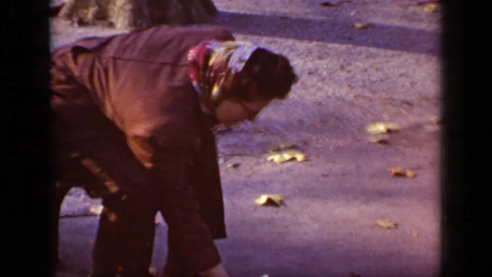 Clearer 1948 New York frame showing an older woman in a brown coat and scarf bending toward a squirrel on a leaf-strewn paved surface.