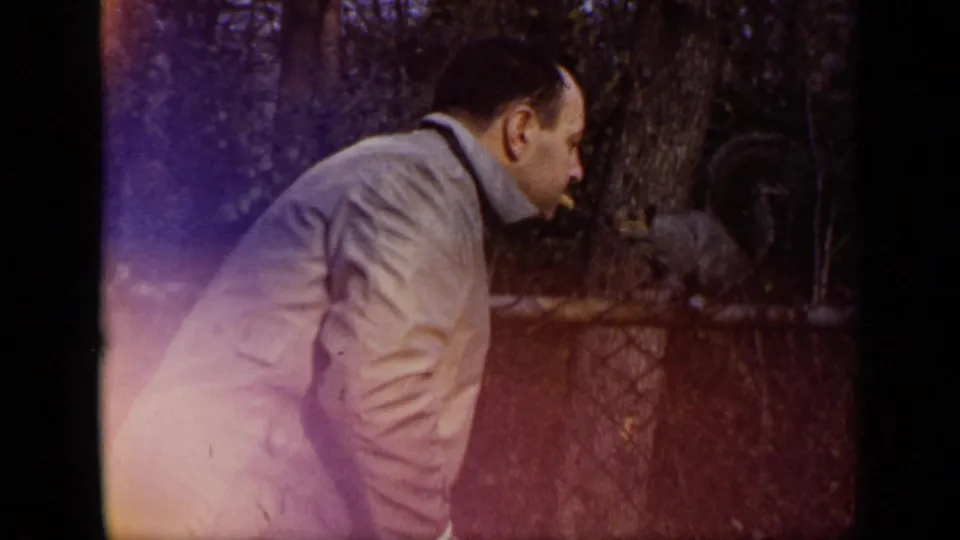 A man in a pale jacket leans toward a squirrel on a fence in wooded 1948 New York home-movie footage.
