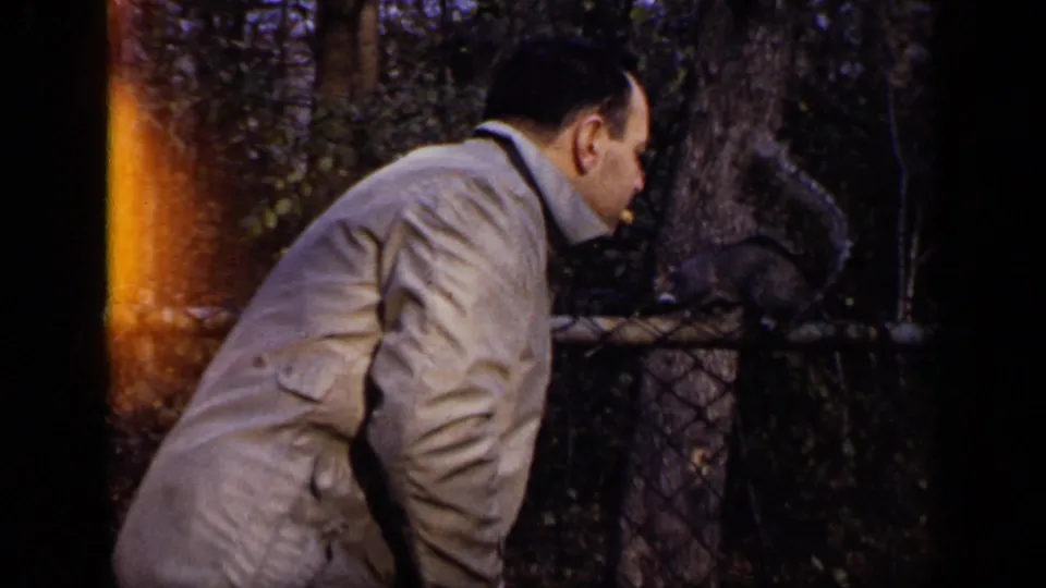 The man remains bent forward, watching a squirrel on the chain-link fence in a shaded New York outdoor setting.