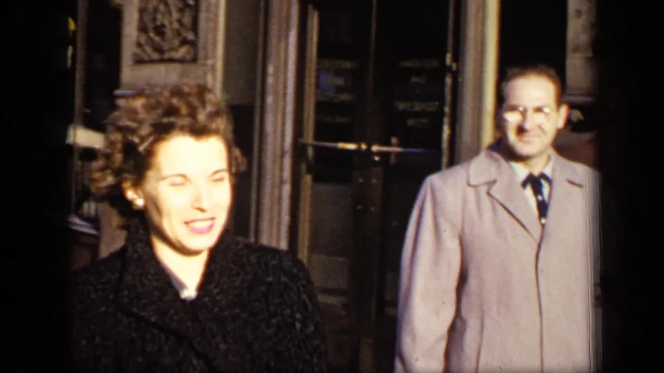 In a 1948 New York home-movie frame, a woman in a dark coat stands close to the camera near a building entrance.