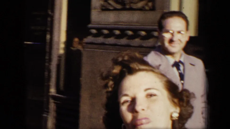 A smiling man in glasses and a light overcoat stands by a dark building entrance in the 1948 New York street footage.