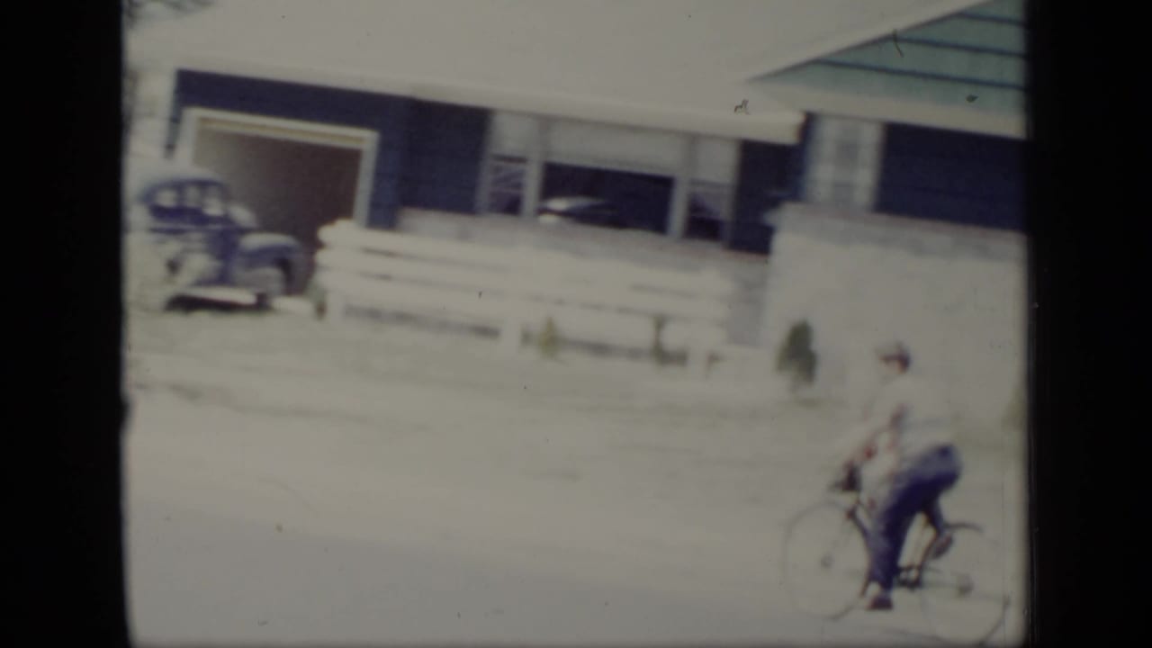Two young riders on a bicycle pass a house front and white fence.