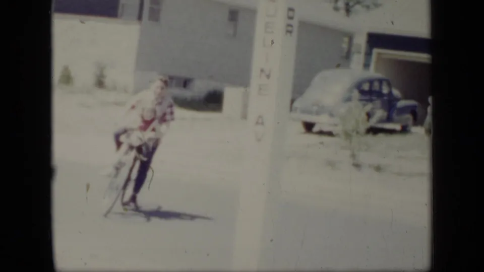 A close, soft 8mm frame shows two young riders leaning together on a bicycle, with a rounded parked car and residential exterior behind them.