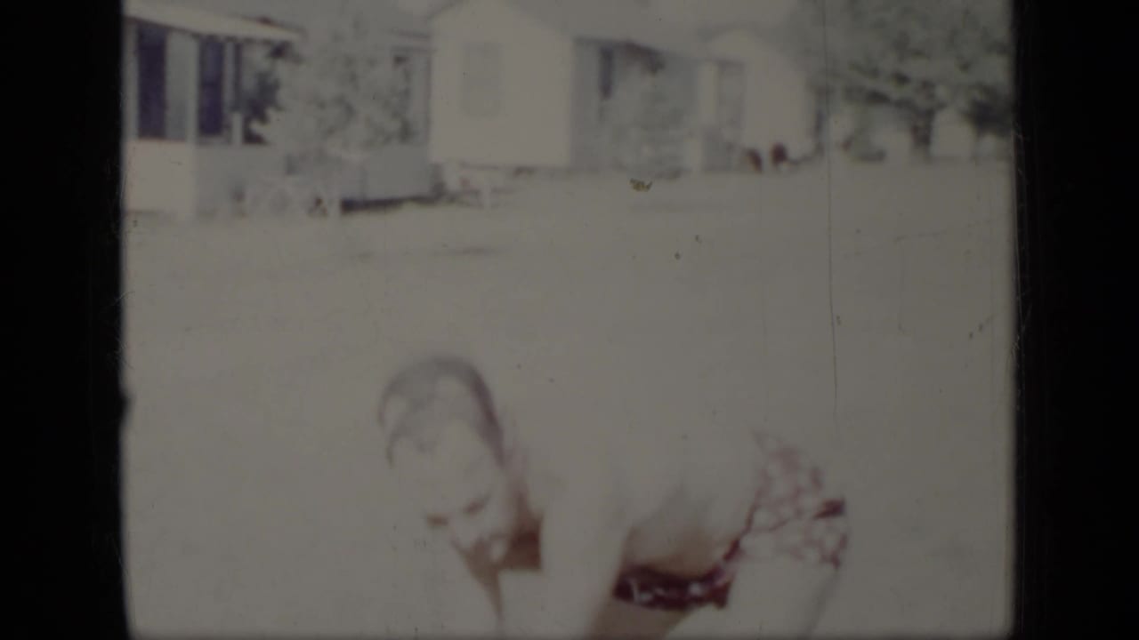 Man in patterned swim trunks bending in a bright yard near a house.