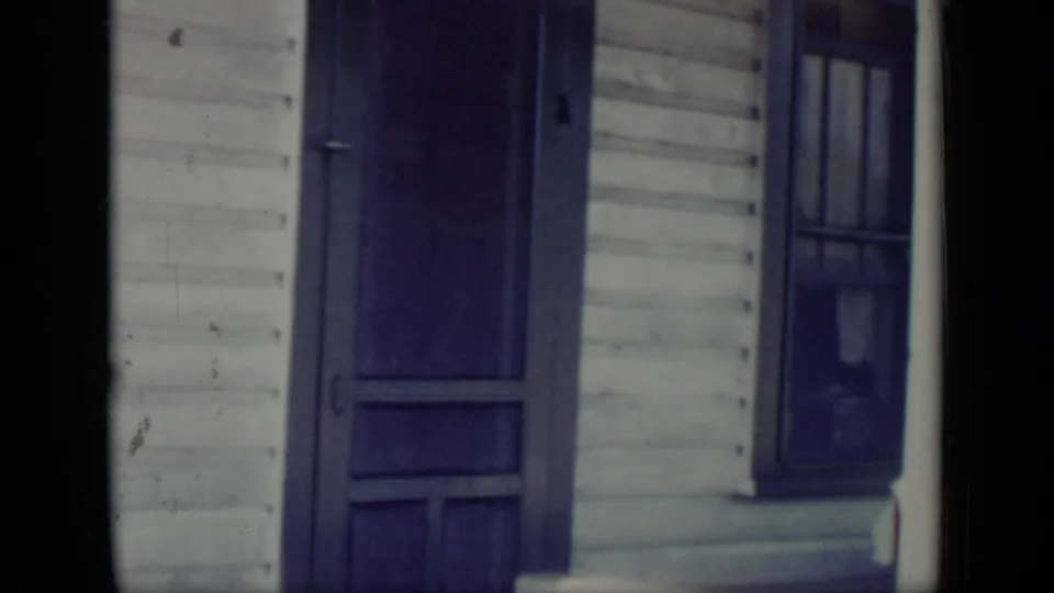 The camera frames a white clapboard house in New York, with a dark screen door and adjacent window visible in soft 8mm color.