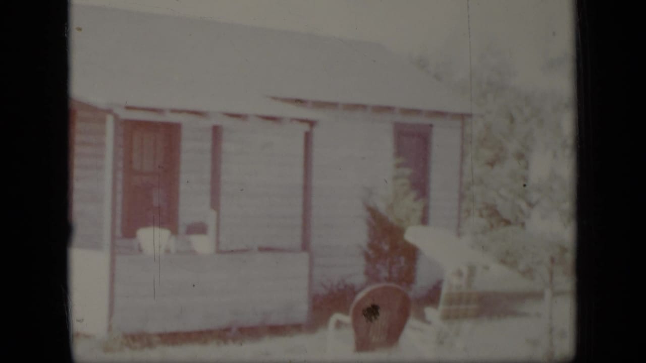 1948 New York 8mm frame showing a white clapboard house exterior with a covered porch, dark doorway, small tree, and chair.