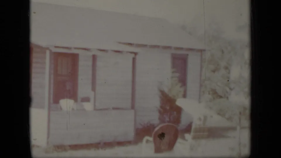1948 New York 8mm frame showing a white clapboard house exterior with a covered porch, dark doorway, small tree, and chair.