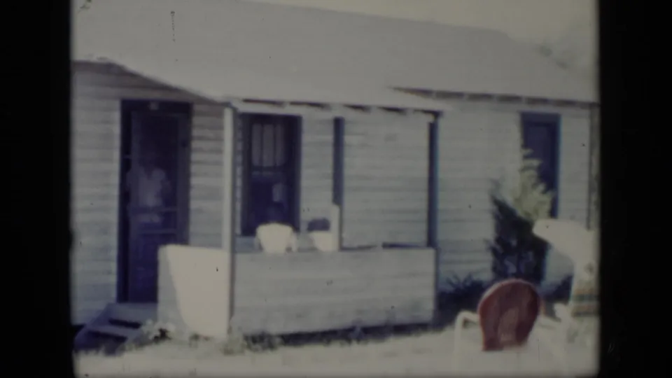 1948 New York home-movie frame with a man standing inside an open doorway beside a small covered porch.