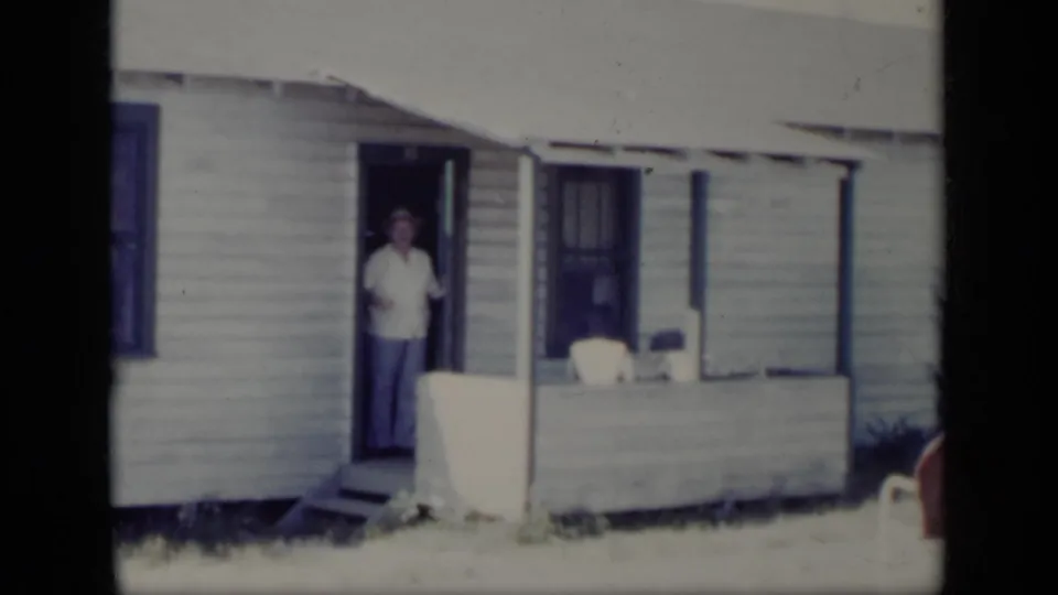 1948 New York 8mm frame showing a man in a light shirt and hat standing in the open doorway of a clapboard house.