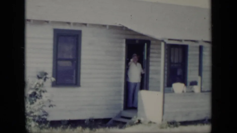 1948 New York home-movie frame of a man raising his hand while standing in the doorway of a small white house.