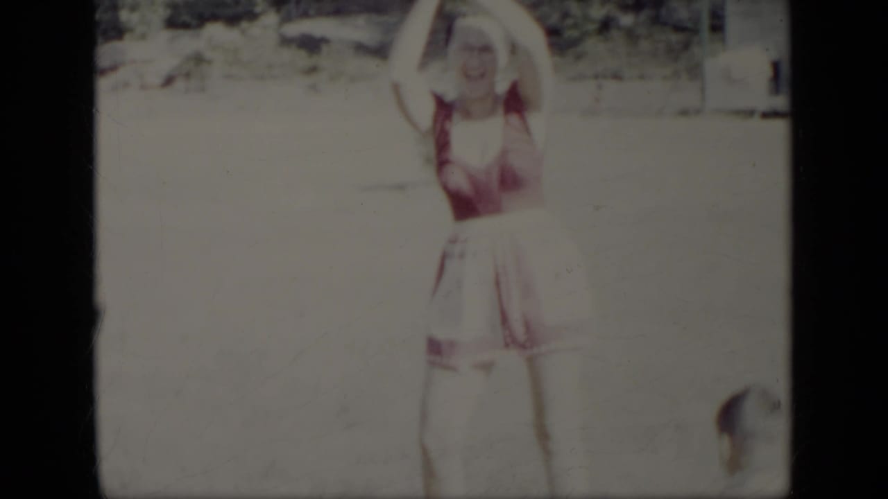 In this 1948 New York home-movie frame, a woman in a red-and-white swimsuit stands in a sunny yard with her arms raised.