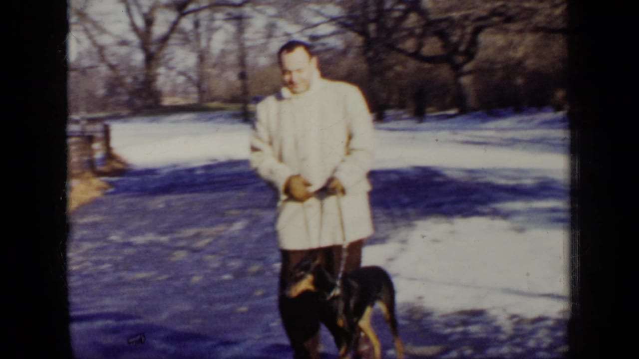 Man in a light winter coat stands with a Doberman on a snowy park path.