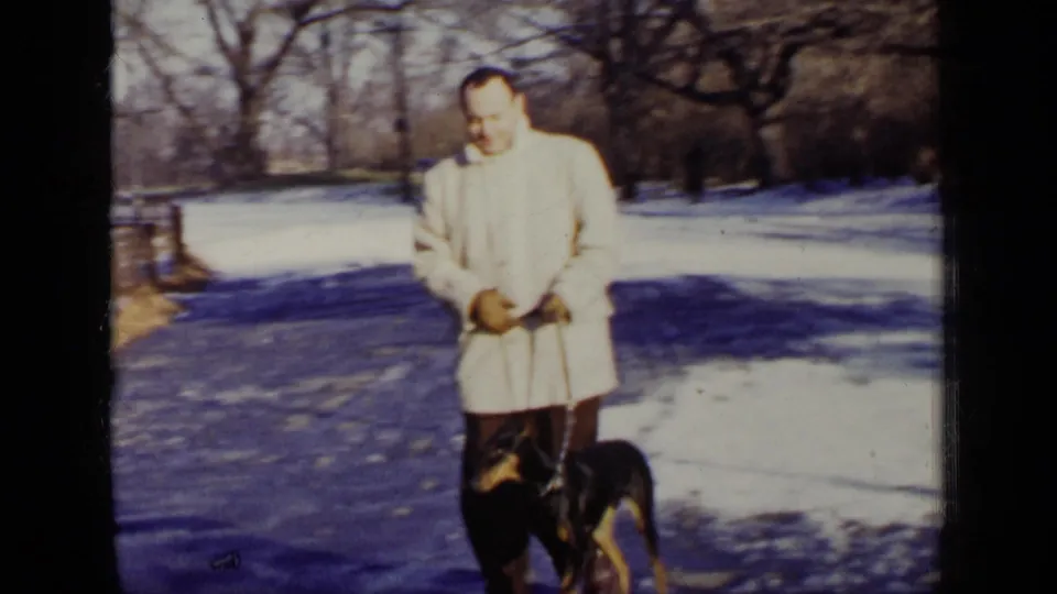 In a 1948 New York home-movie frame, a man in a light coat holds a Doberman on leash beside snow and bare trees.