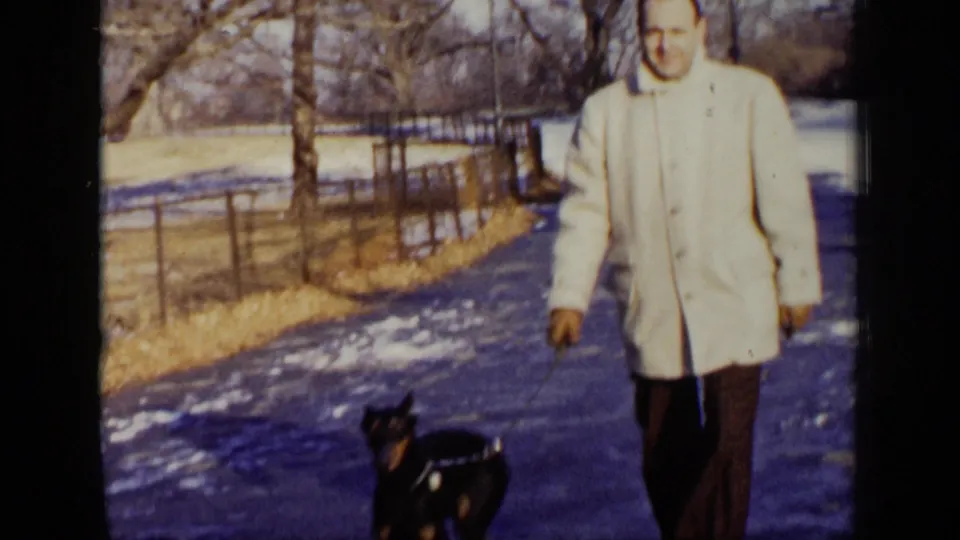 The man walks the Doberman along a New York park path, with snow patches, bare trees, and a wire fence behind them.