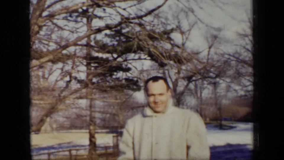 A closer home-movie view shows the man smiling in a light winter coat, with bare trees and snow-dusted park ground behind him.